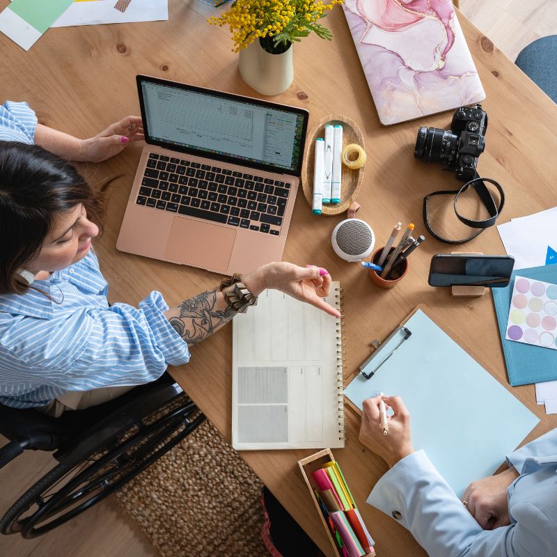 Dos personas trabajando en un oficina compartiendo información con un ordenador y varios documentos y material de oficina sobre la mesa.